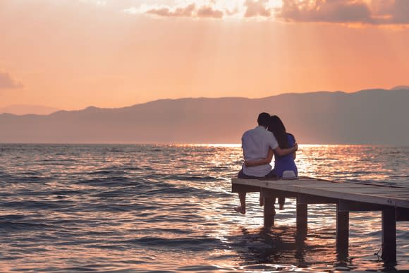 A couple sits on the end of a short dock on the water facing a sunset with their arms around each other.