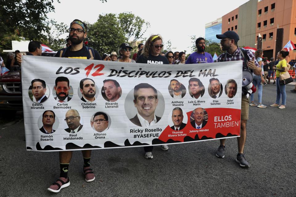 Protesters hold a banner featuring resigned Gov. Ricardo Rosselló, center, amid other politicians that reads in Spanish "The 12 disciples of evil. Them too." as they demand the resignation of Justice Secretary Wanda Vazquez outside the Department of Justice in San Juan, Puerto Rico, Monday, July 29, 2019. Less than four days before Gov. Ricardo Rosselló steps down, no one knows who will take his place and his constitutional successor Wanda Vázquez said Sunday that she didn't want the job. (AP Photo/Brandon Cruz González)