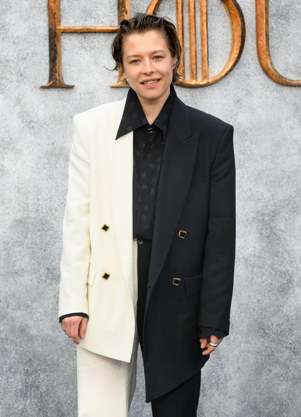 Emma D'arcy poses on the red carpet, wearing a distinctive asymmetric half white, half black suit jacket over a black shirt