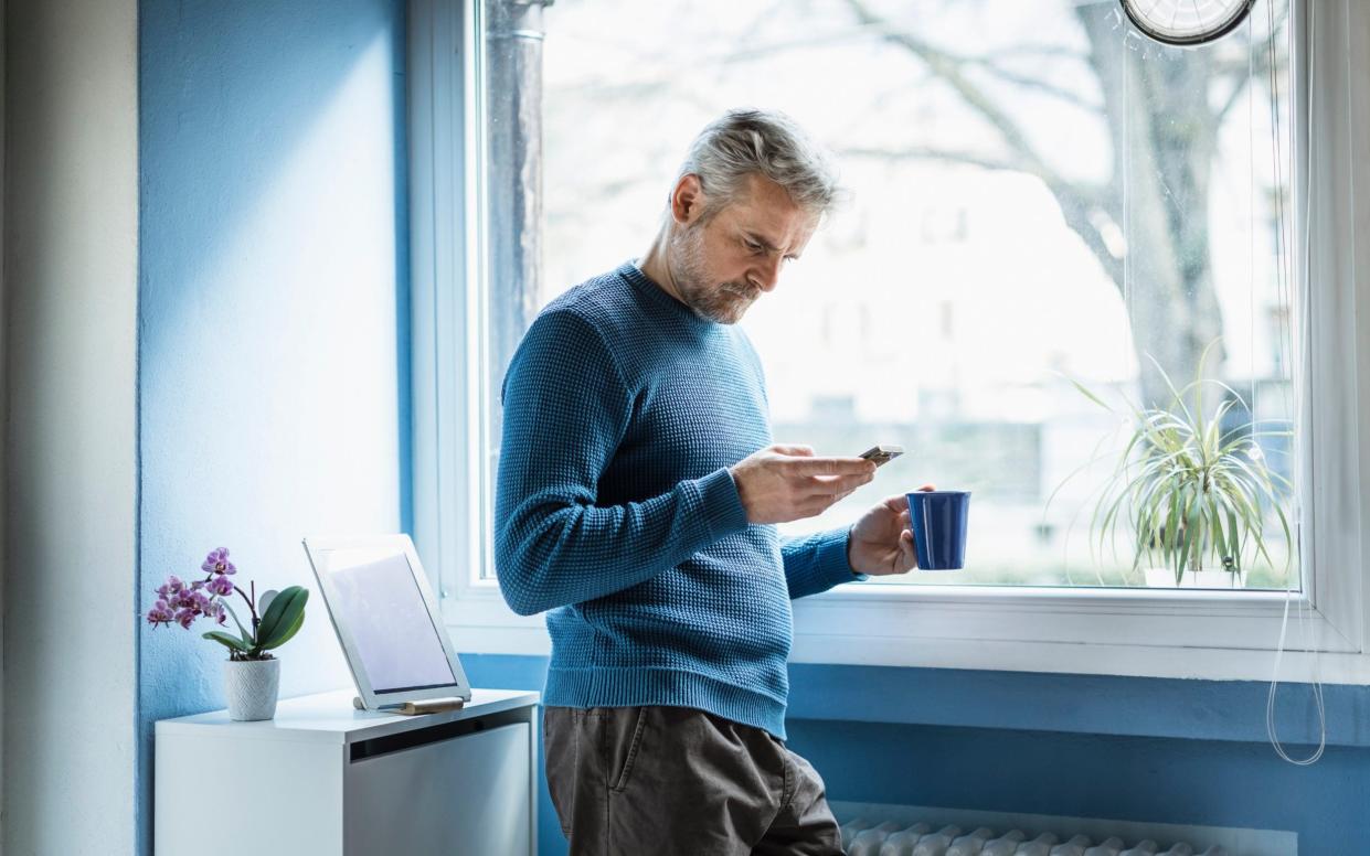 Mature man standing with coffee mug standing in living room looking at smartphone