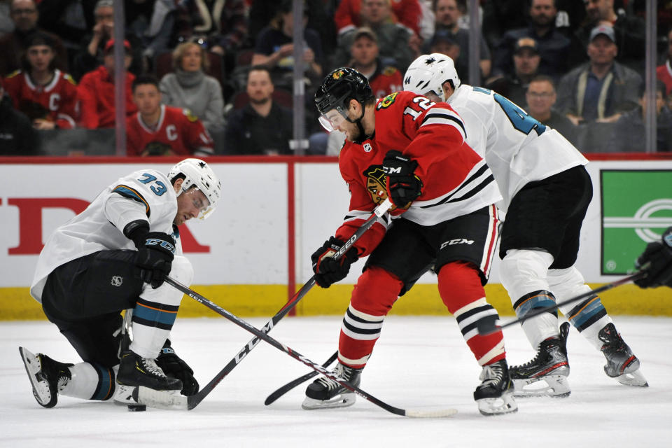 Chicago Blackhawks' Alex DeBrincat (12) battles San Jose Sharks' Noah Gregor (73) and Marc-Edouard Vlasic (44) for a loose puck during the second period of an NHL hockey game Wednesday, March 11, 2020, in Chicago. (AP Photo/Paul Beaty)