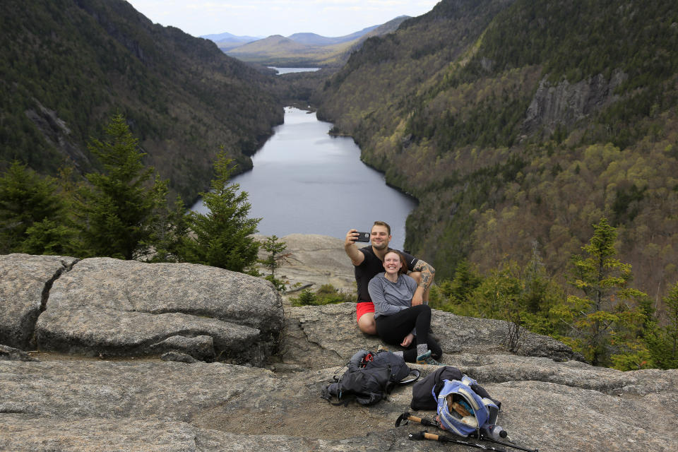 Sidney Gleason, right, and Joe Gorsuch, both of Syracuse, take a selfie with Lower Ausable Lake in the background, while taking a break at Indian Head summit inside the Adirondack Mountain Reserve, Saturday, May 15, 2021, near St. Huberts, N.Y. A free reservation system went online recently to control growing number of visitors packing the parking lot and tramping on the trails through the private land of the Adirondack Mountain Reserve. The increasingly common requirements, in effect from Maui to Maine, offer a trade-off to visitors, sacrificing spontaneity and ease of access for benefits like guaranteed parking spots and more elbow room in the woods. (AP Photo/Julie Jacobson)
