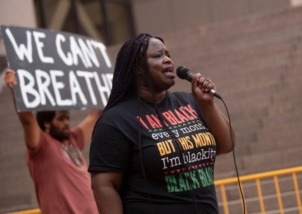 nekima levy armstrong speaks into a microphone she holds in one hand as she stands outside, she wears a black tshirt with writing on it, a man stands behind to the left with a protest sign