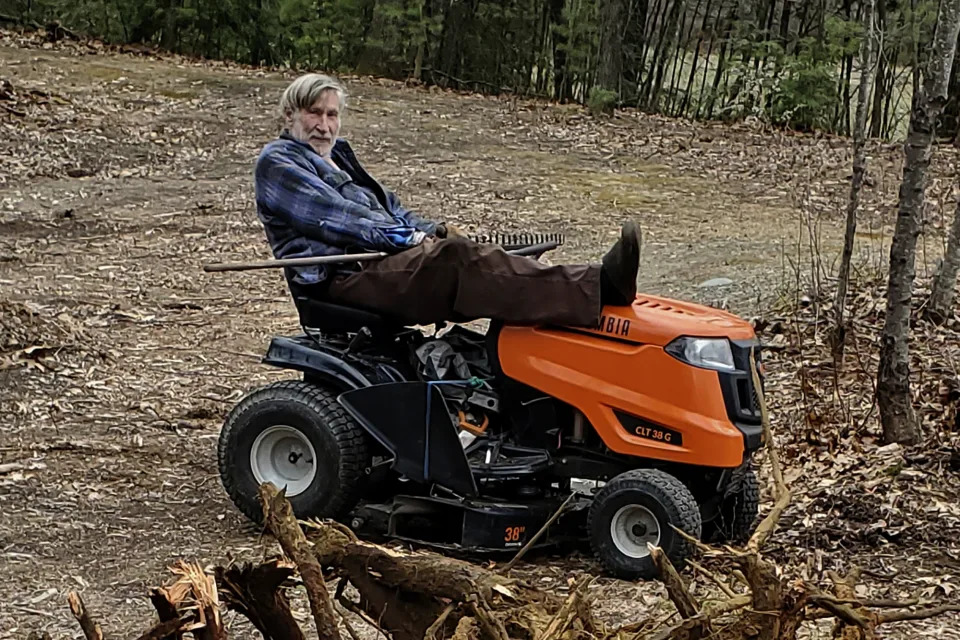 In this photo provided by Ed Smith, Geoffrey Holt rests his leg on top of his riding mower in Hinsdale, N.H., on April 4, 2020. Holt left the town of Hinsdale nearly $4 million when he died last June. (Ed Smith via AP)