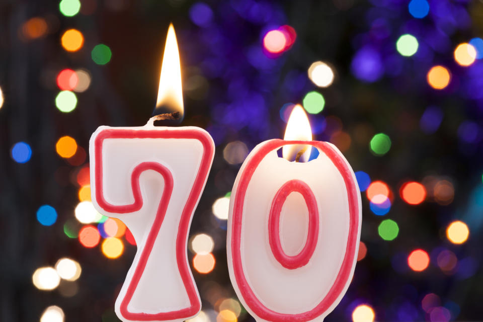 Two lit candles are shown, in front of blurry colored lights, one candle is the number seven and the other is a zero, together representing the number seventy.