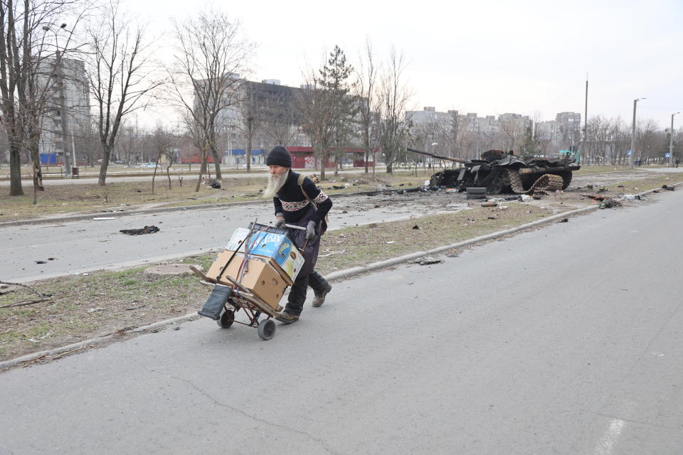 A man wheeling boxes on a dolly walks past a damaged tank by the side of the road.