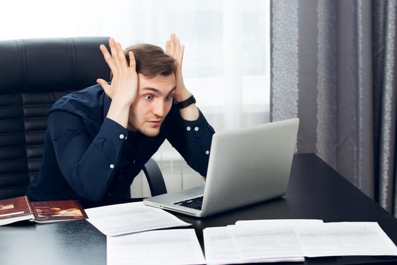 A young man stares at his laptop in shocked confusion, hands pressing against his temples and eyes wide open.