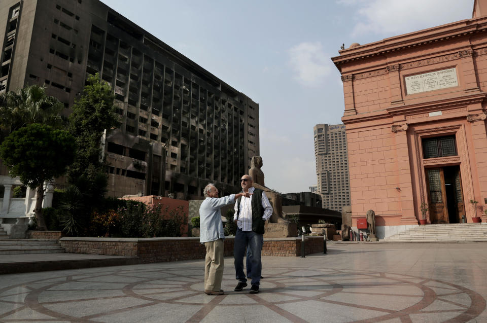 In this Wednesday, Oct. 30, 2013 photo, tourists chat with the nearby burned headquarters of the former ruling National Democratic Party, background left, in the Egyptian Museum near Tahrir Square in Cairo, Egypt. The 111-year-old museum, a treasure trove of pharaonic antiquities, has long been one of the centerpieces of tourism to Egypt. But the constant instability since the 2011 uprising that toppled autocrat Hosni Mubarak has dried up tourism to the country, slashing a key source of revenue. Moreover, political backbiting and attempts to stop corruption have had a knock-on effect of bringing a de facto ban on sending antiquities on tours to museums abroad, cutting off what was once a major source of funding for the museum. (AP Photo/Nariman El-Mofty)