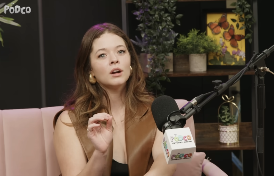 Sasha Pieterse is speaking into a microphone during an episode of Pretty Little Liars: True Crime, seated in a studio with plants in the background