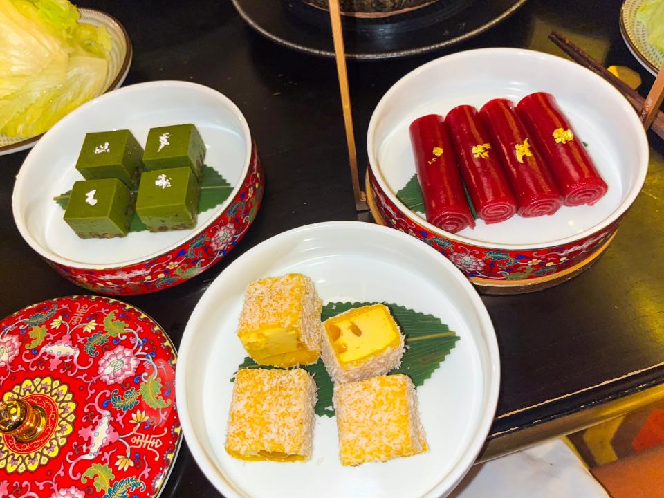 A black table filled with multicolored bowls with white interiors filled with desserts. The bowls are filled with square green pastries, square yellow pastries, and red roll-up pastries.