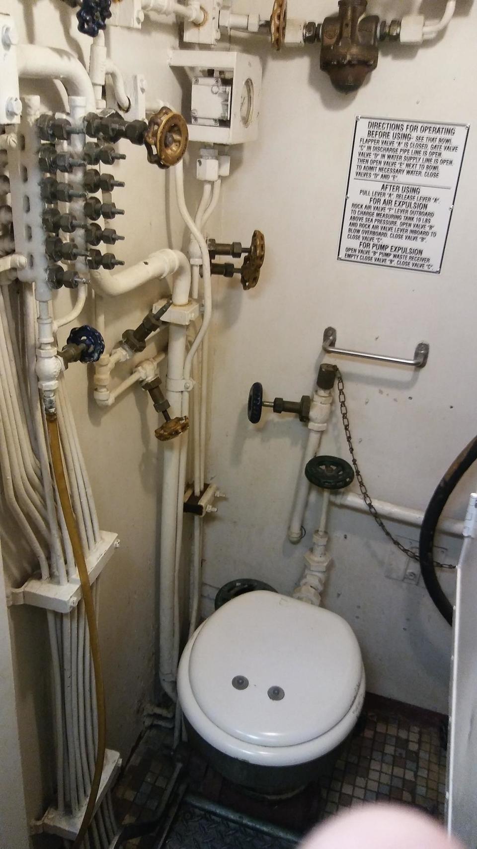 A toilet bowl surrounded by pipes and chains and cords and valves and knobs, with a sign with lots of text labeled, "Directions for operating: before and after using"