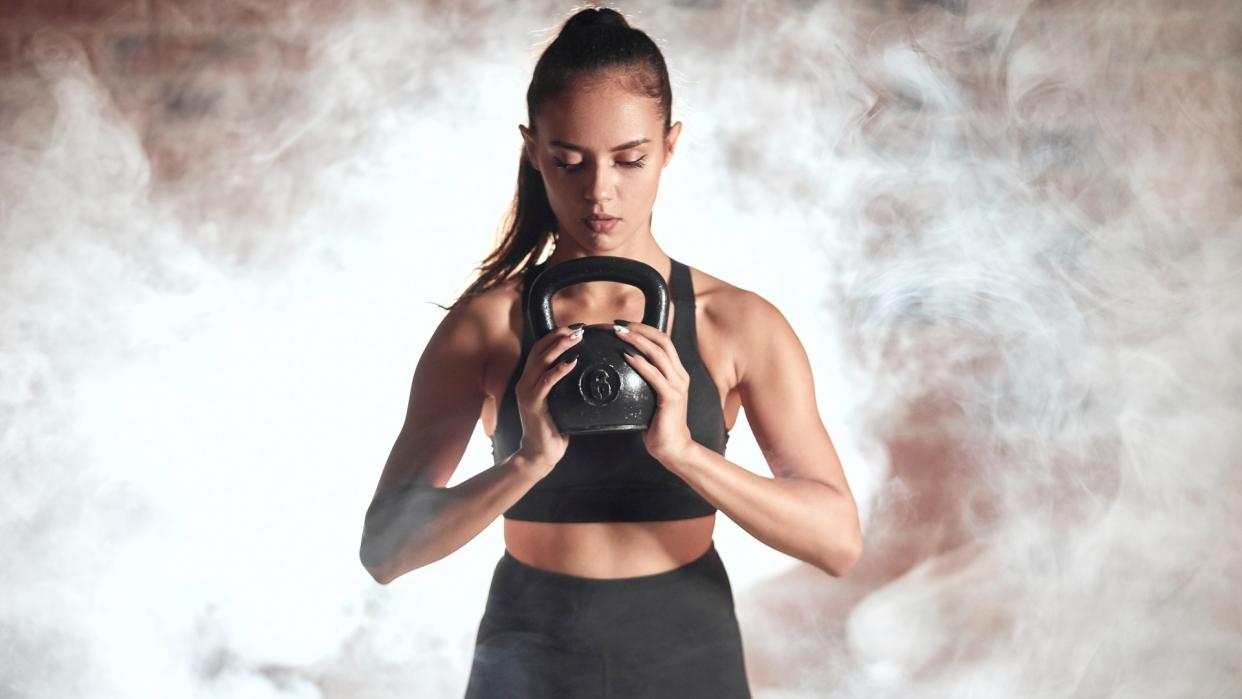  Woman holding a kettlebell in both hands with smoke behind her during kettlebell workout. 