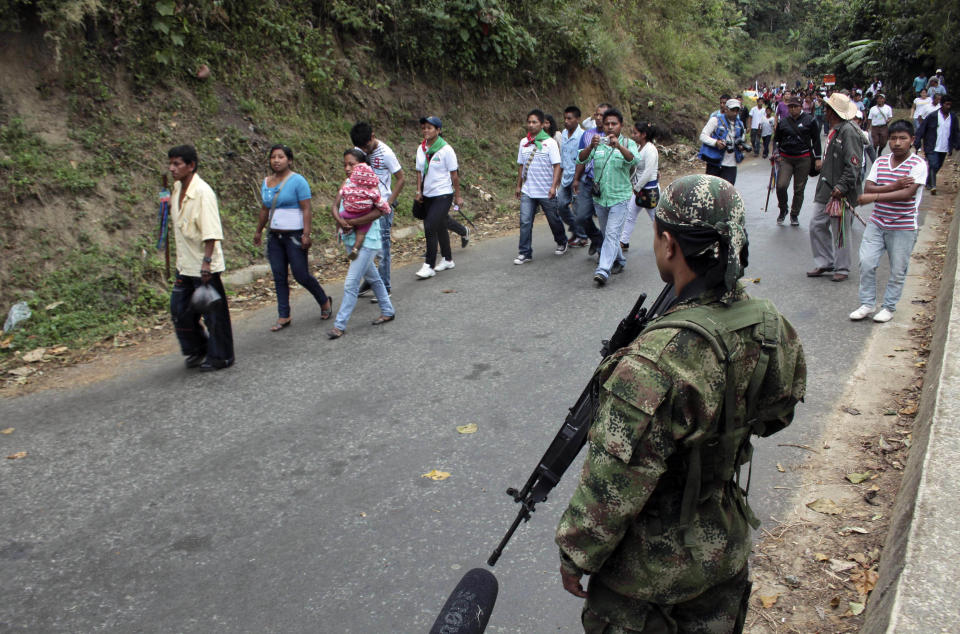 Indians make their way past a rebel of the Revolutionary Armed Forces of Colombia, FARC, front, on the outskirts of Toribio, southern Colombia, Wednesday, July 11, 2012. Rebels set up a roadblock on a road leading to Toribio while Colombia's President Juan Manuel Santos was holding a meeting with cabinet members and local authorities in the church of the town, that was attacked by guerrillas last week. (AP Photo/Juan Bautista Diaz)