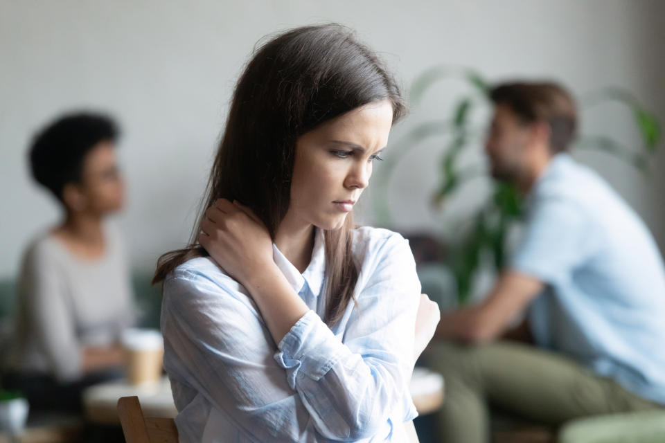 Upset young woman sitting alone in cafe offended at diverse friends, depressed female feeling outsider, depressed, jealous, suffering from low self-esteem, discrimination, hugging herself