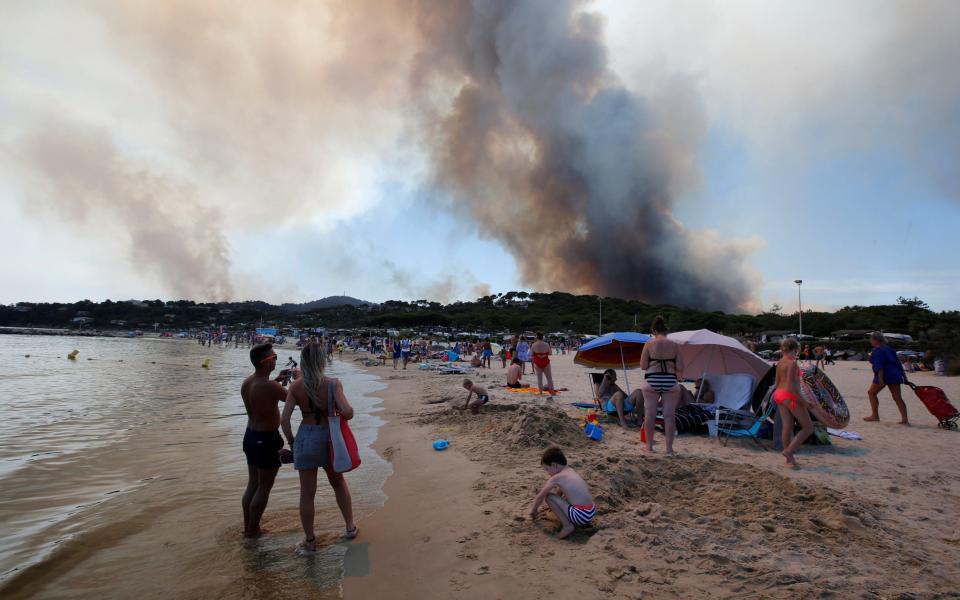 Tourists stand on the beach and watch as smoke fills the sky above a burning hillside in Bormes-les-Mimosas