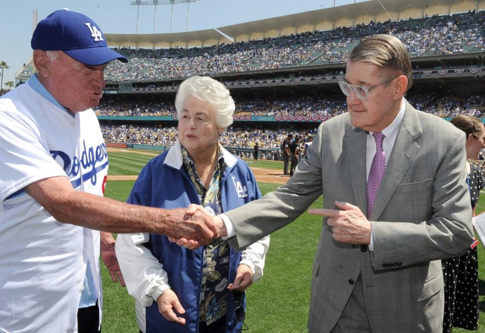 Two men shake hands while a woman watches as people fill the stands behind them at a baseball stadium.