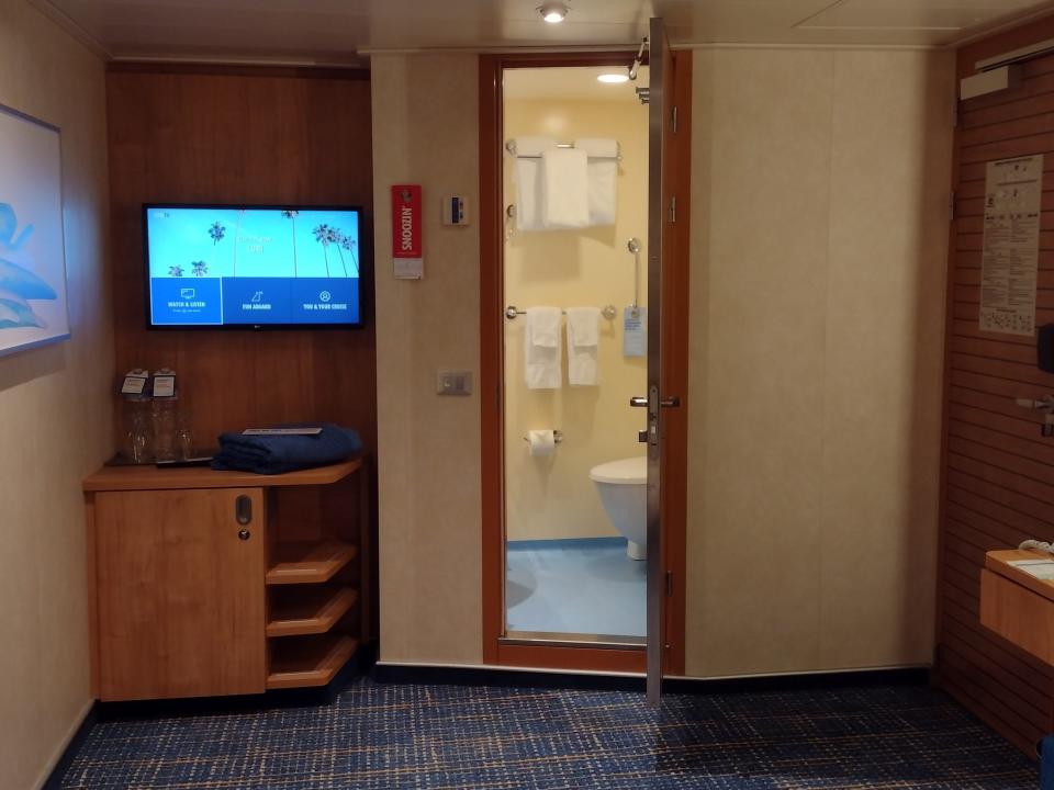 A cruise ship room with the view facing the TV, bathroom, and door.
