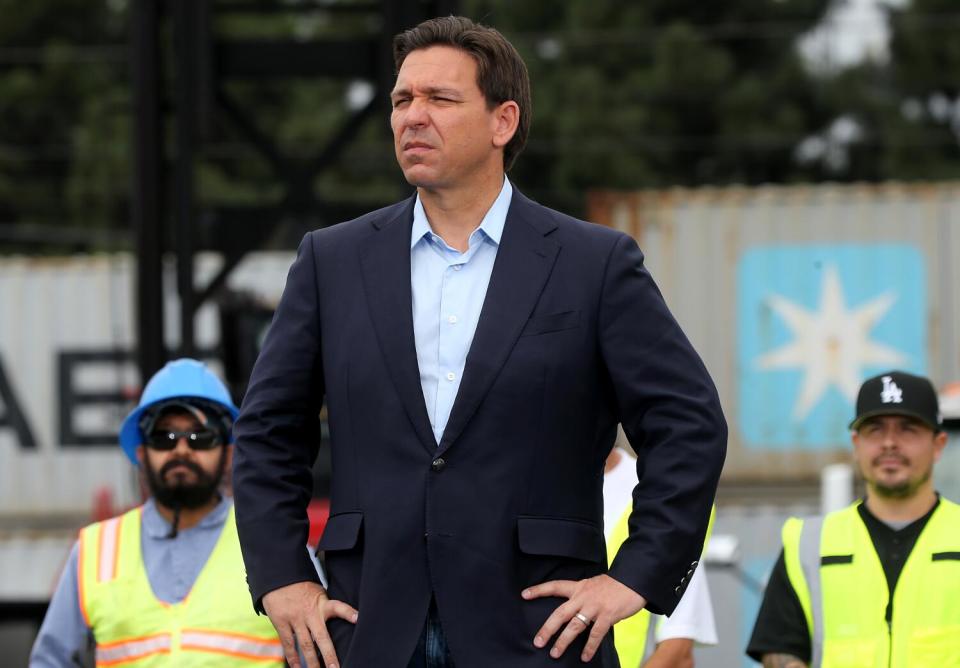 A man with dark hair, in blue shirt and dark jacket, stands with hands on hips