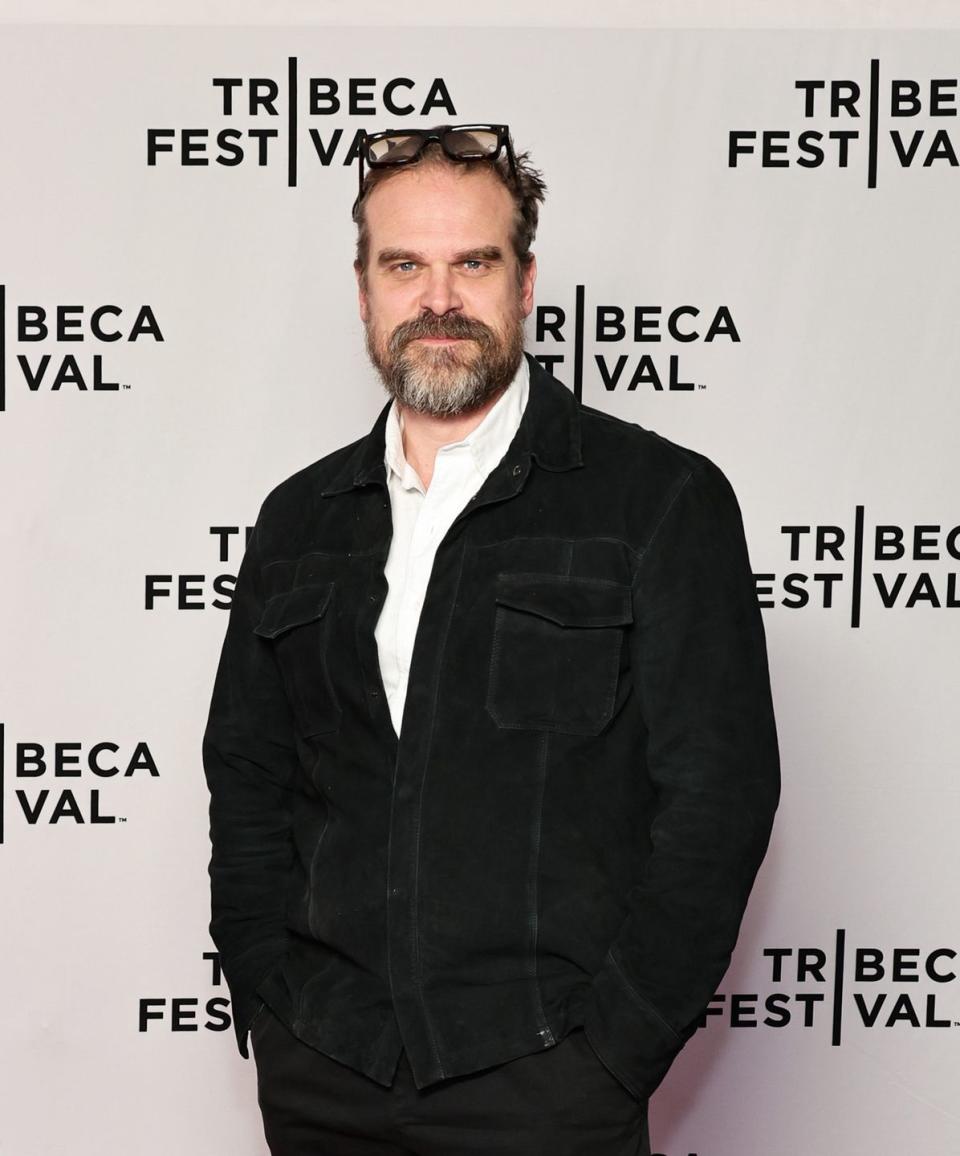 david harbour, a man with short brown hair and a beard stands looking at the camera with a straight face and hands in his trouser pockets, he wears glasses on top of his head, a white shirt, black trousers and black jacket