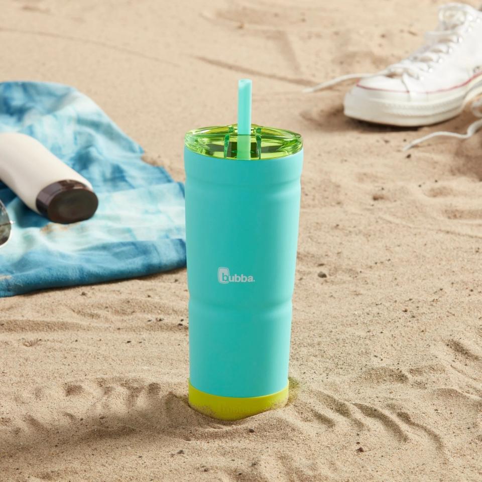 A Bubba brand tumbler on sandy beach with a blue towel and white sneakers nearby