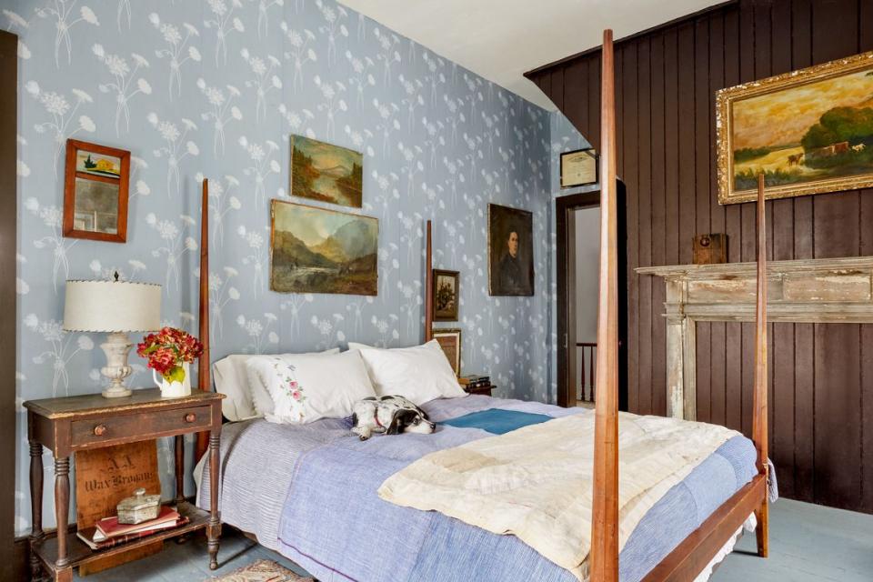 a farmhouse bedroom that has an statement accent wall in a light blue with white flowers wallpaper and theres a pencil post bed with a little black and white dog on it