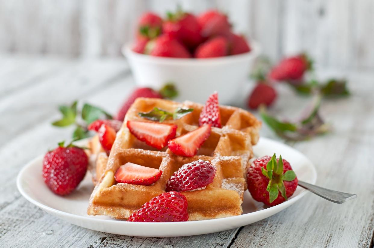 A strawberry waffle with strawberries on a white plate with a fork on a light grey wooden table with a blurred background of a white bowl of strawberries