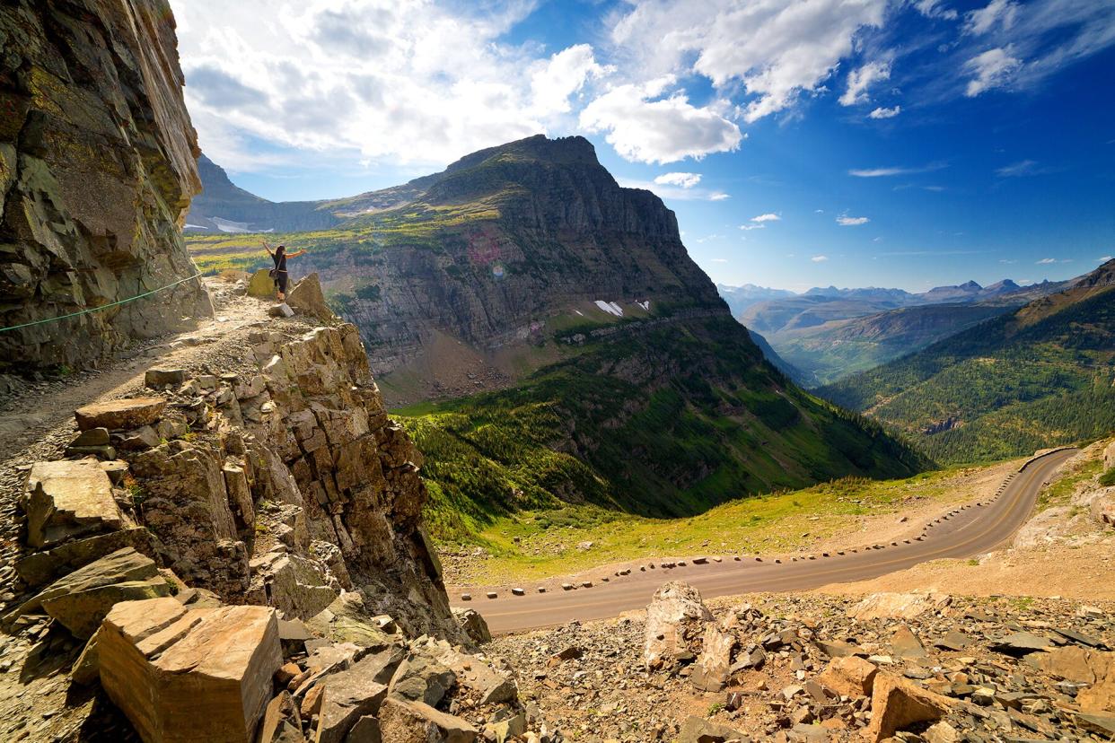 Female hiker on narrow highline trail stands on overlook with arms outstretched, looking down on going-to-the-sun road