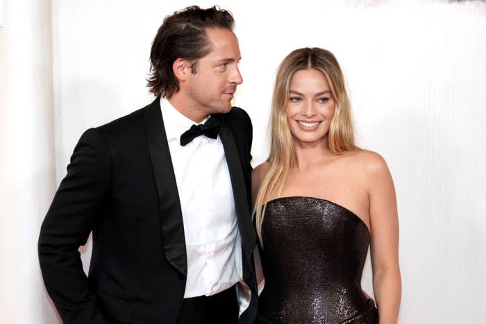 Tom Ackerley and Margot Robbie smile for the camera; Tom is in a black tuxedo, and Margot is in a strapless, glittering black gown