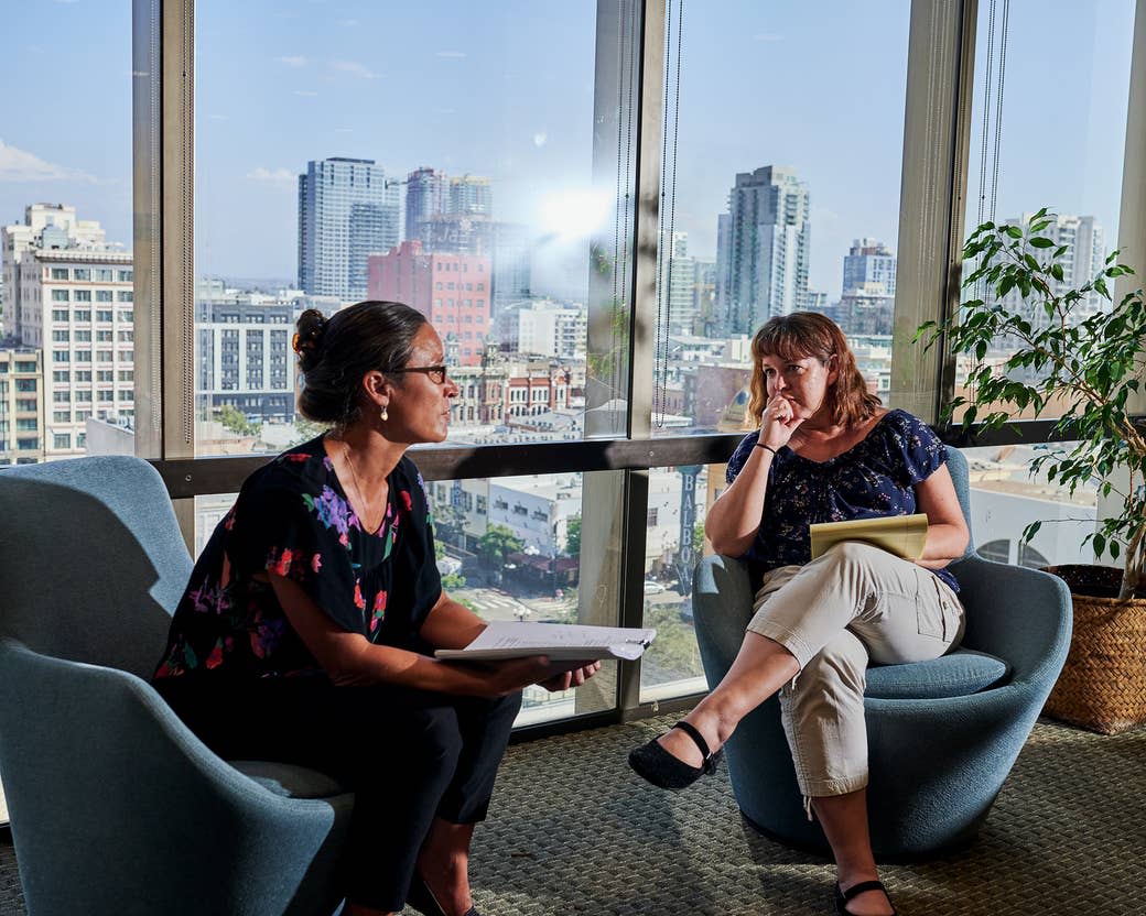 Two women sit next to each other with the city visible in the floor-length windows behind them