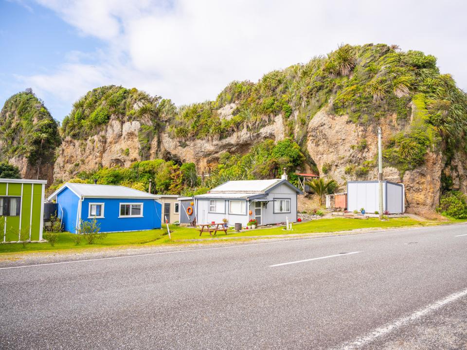 A row of colorful houses next to a road in front of tree-topped rocks.