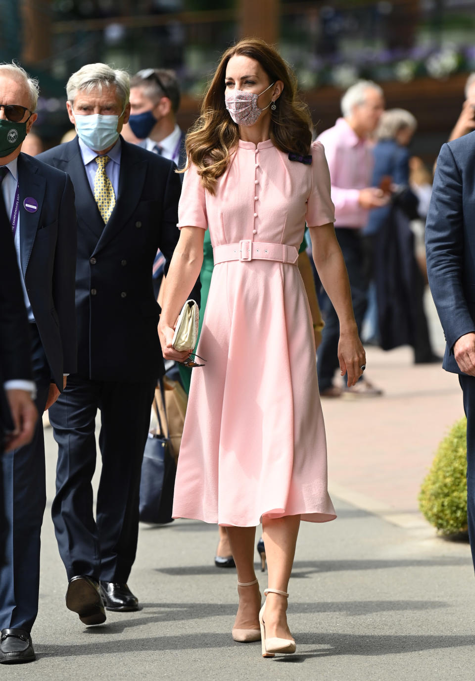 Michael Middleton and Catherine, Duchess of Cambridge attend day 13 of the Wimbledon Tennis Championships wearing masks. Kate wears a pink buttoned dress with a wide pink belt and a skirt below the knee and high heels. Her hair is loose.