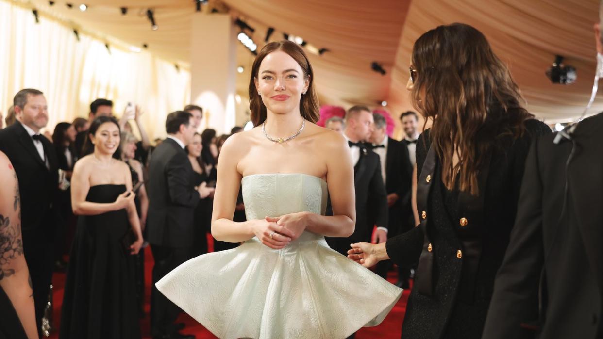 emma stone smiles as she walks on a red carpet in a strapless dreess, several people are in the background