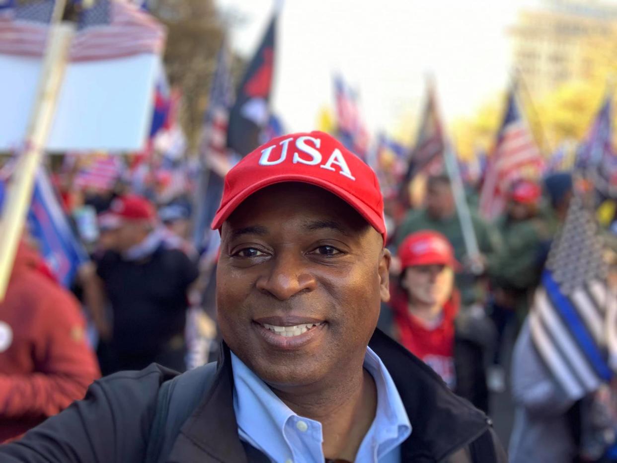 Leon Benjamin, wearing a red baseball cap that says USA takes a selfie in a crowd carrying flags and posters.