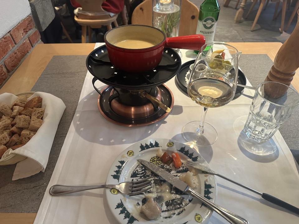 fondue pot full of melted cheese on a restaurant table beside plate of pickles glass of white wine cup of water and basket of bread cubes with a plate holding a cheesy fondue fork