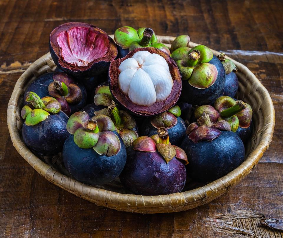 A bowl of mangosteens are in a basket on a wooden table with one having its exterior removed