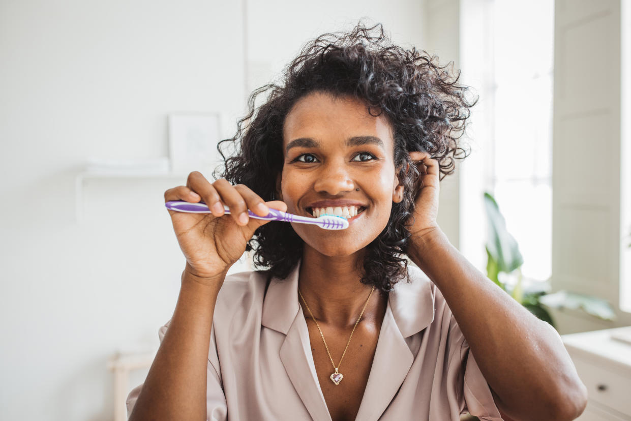 Curly-haired woman brushing her teeth while pushing her hair behind her left ear.