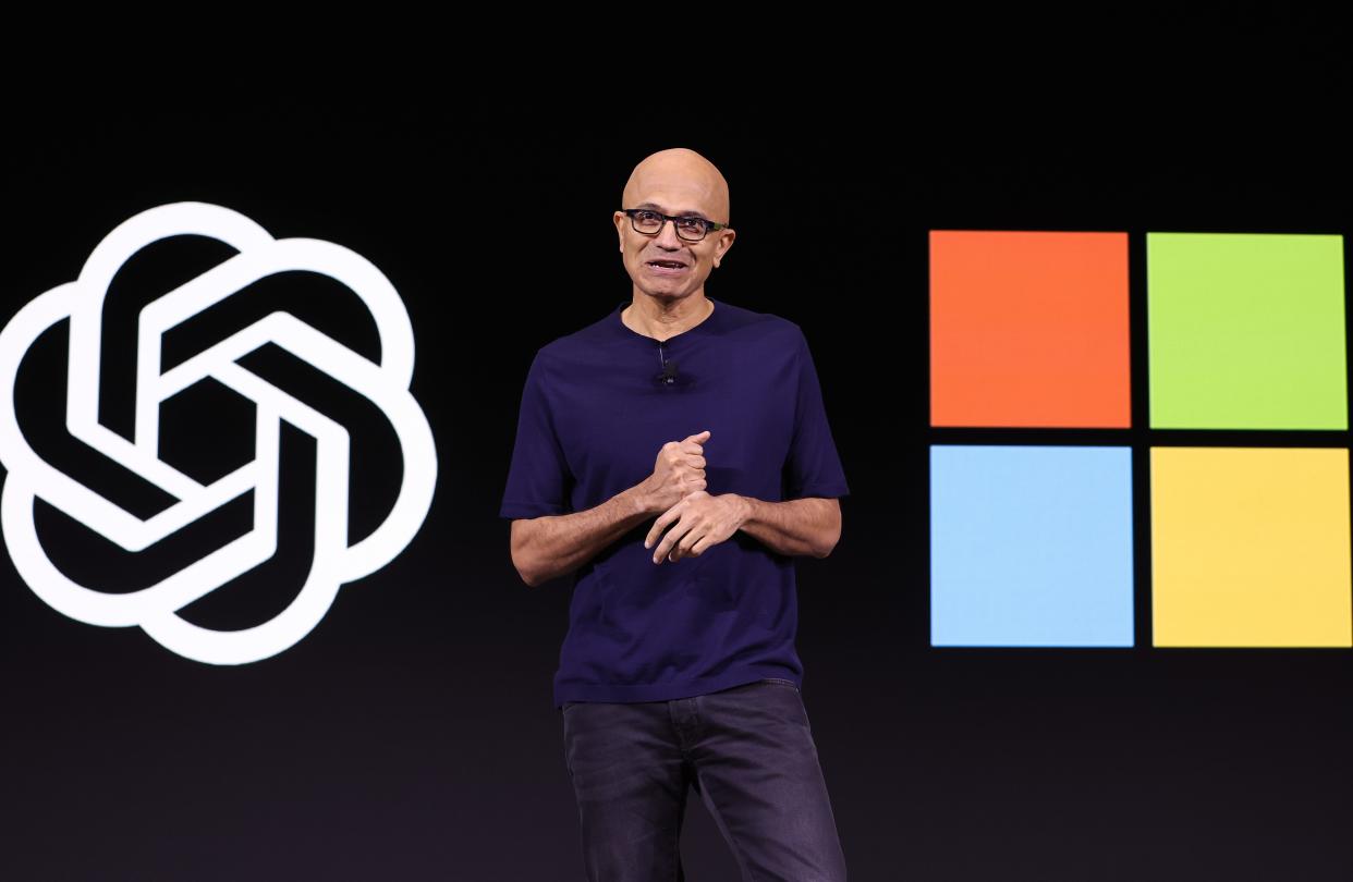 Satya Nadella standing between logos for OpenAI and Microsoft.