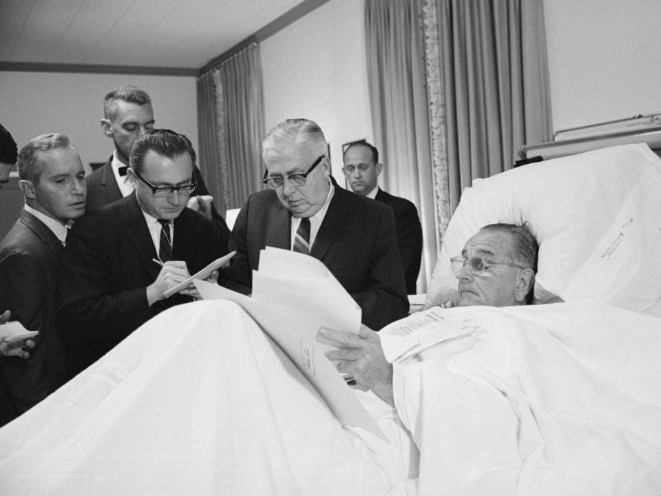 With newsmen standing beside him, then-President Johnson looks over some documents in his hospital bed, November 16th, after undergoing surgery at the Bethesda Naval Hospital here. The facility merged with Walter Reed.