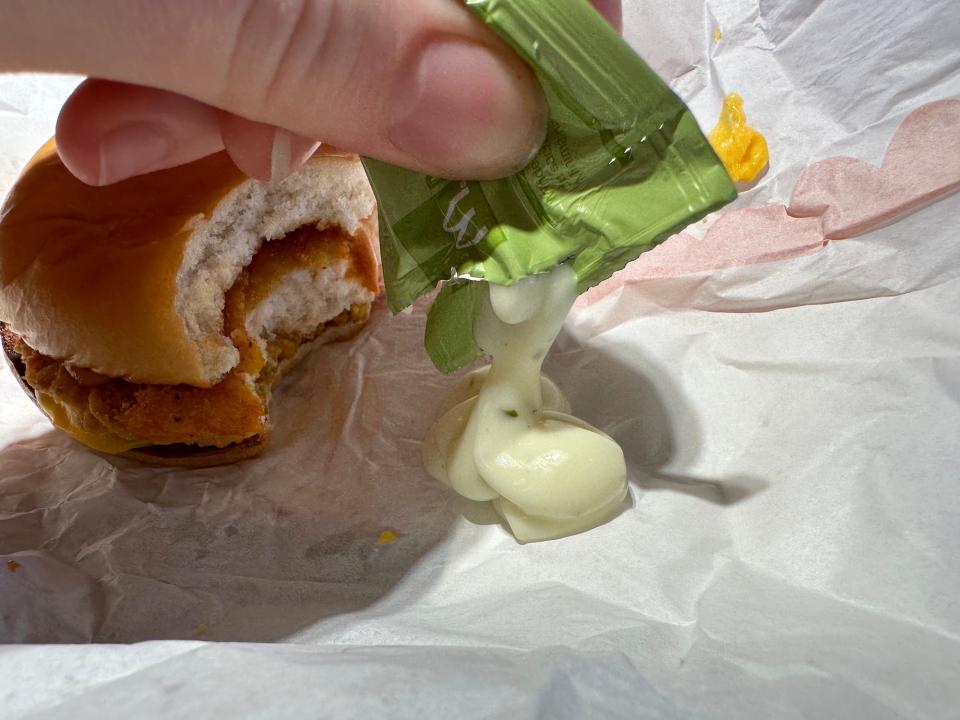 fingers squeeze shiny white sauce out of a green packet onto a white paper wrapping next to a chicken sandwich