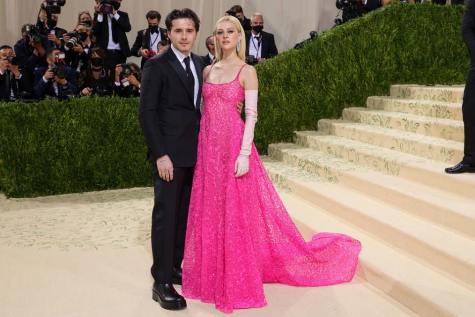 Brooklyn Beckham wears a dark suit and Nicola Peltz wears a thin strap lace brightly colored gown with elbow length gloves