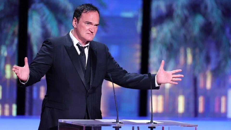 Quentin Tarantino, a light-skinned man with short black hair, wears a tuxedo and speaks in front of a dais with two microphones coming up from it. He extends his arms out, gesturing, in front of a background of a faux skyline with palm trees.