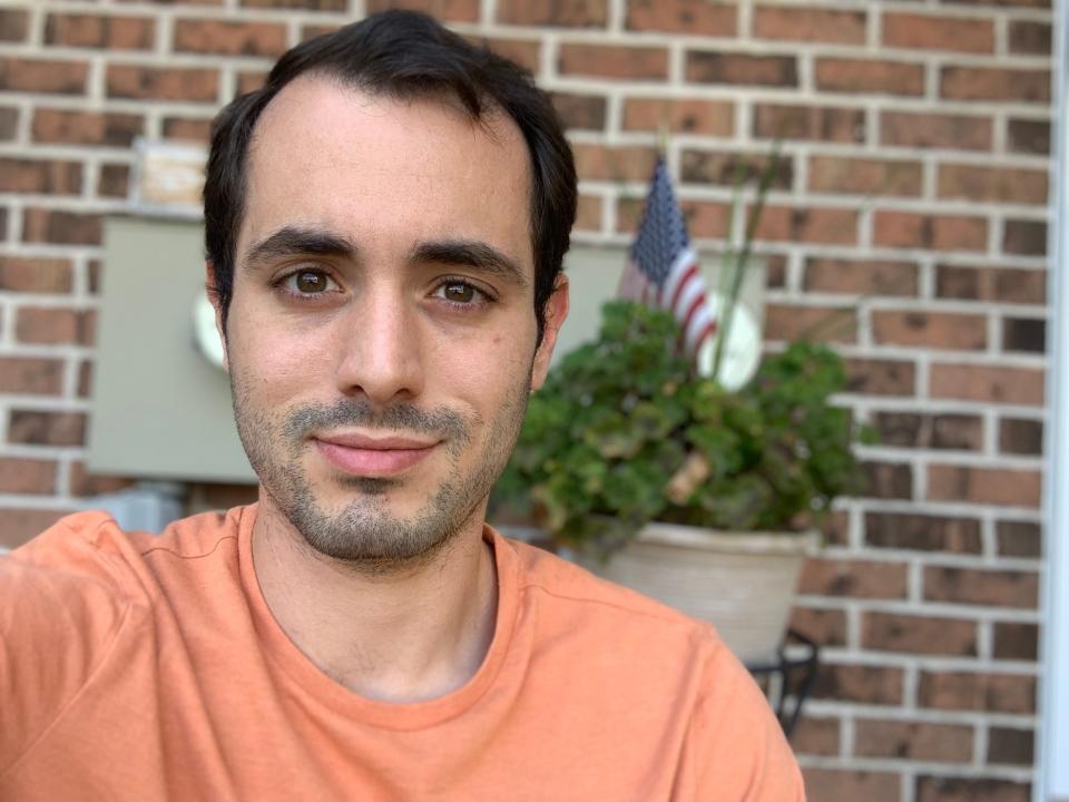 A man with black hair and a light orange t-shirt looks directly into the camera in front of a brick wall.