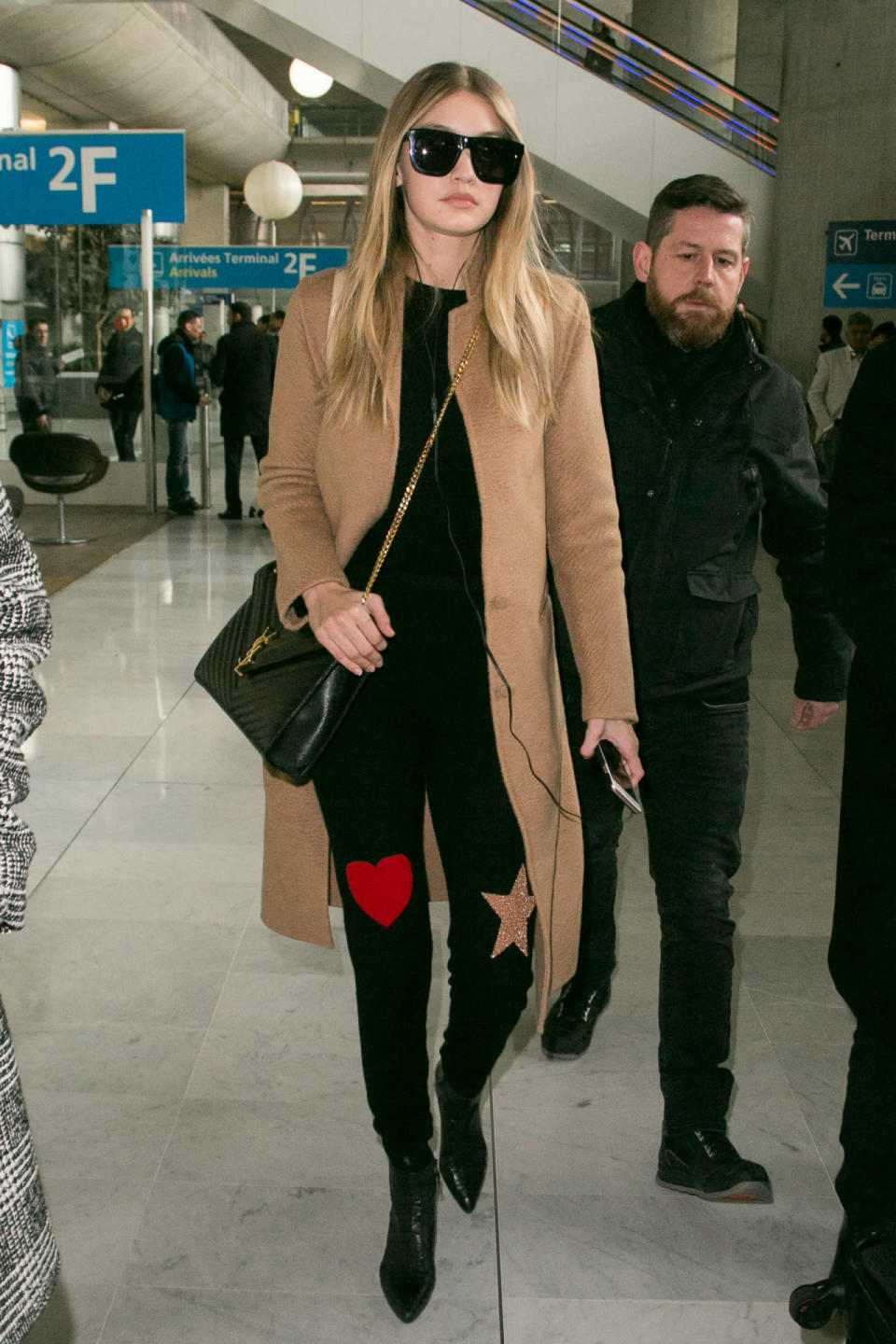 Gigi Hadid wears black skinny jeans with patches and a camel coat as she arrives at Charles-de-Gaulle airport on January 19, 2016 in Paris, France.