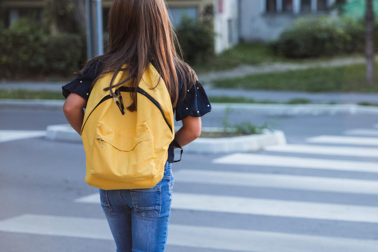 A back view of a long-haired schoolgirl in jeans and a short-sleeve top walking while wearing a yellow backpack.