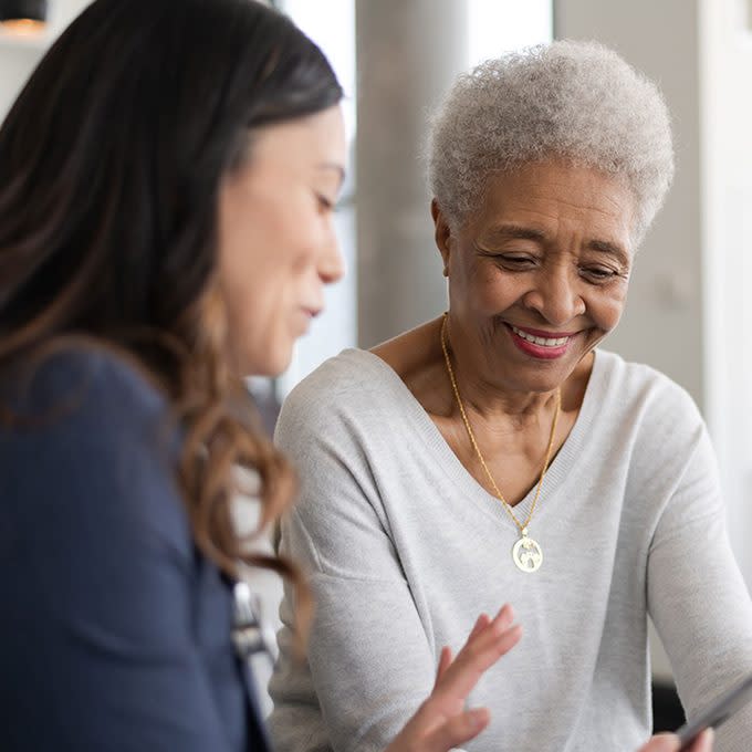 A young doctor and an elderly patient are indoors at the woman's home. The doctor is talking to the woman while holding a tablet computer. They're both smiling.