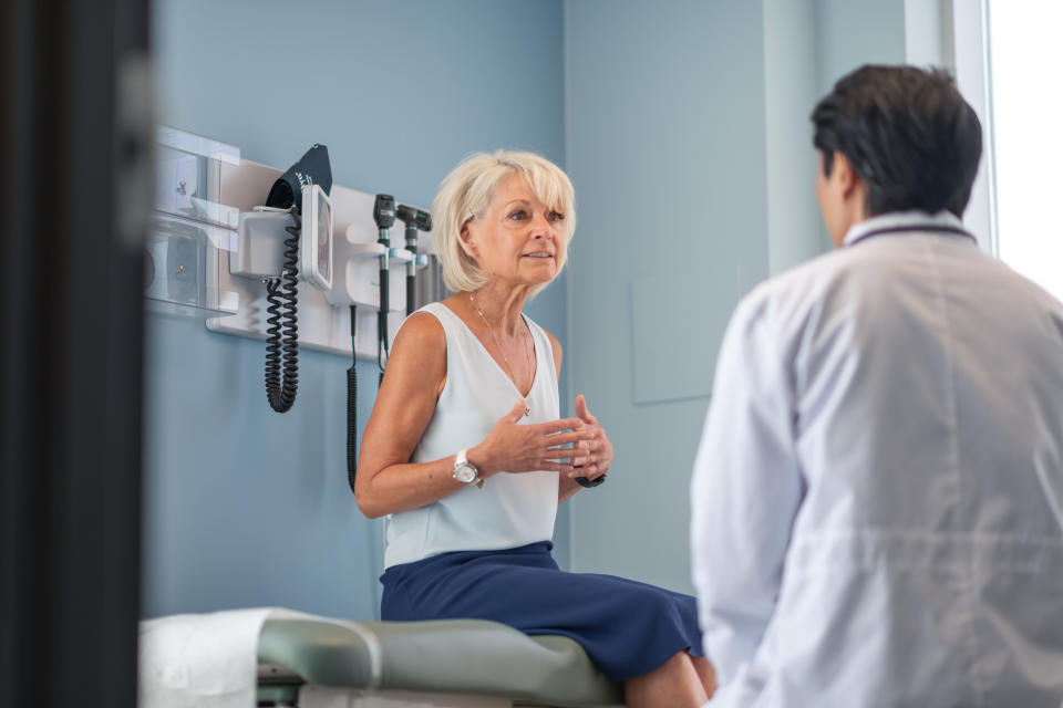 A mature adult woman is at a routine medical appointment. Her healthcare provider is a Korean man. The patient is sitting on an examination table in a clinic. She is explaining her medical history. The kind doctor is listening intently.