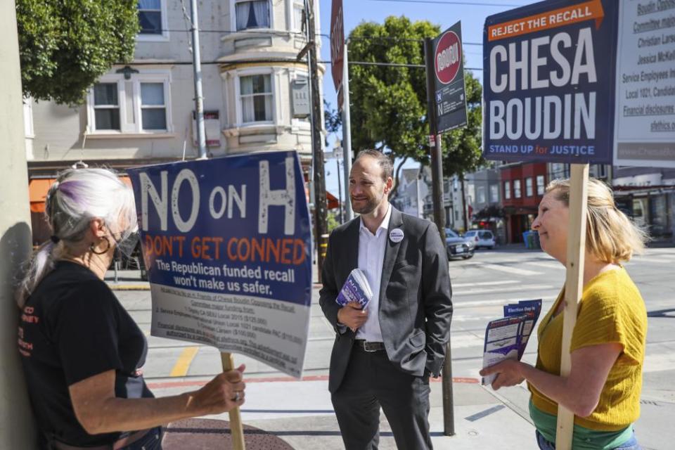 A man in a suit talks to two women on the streets of San Francisco. One holds a sign reading ‘Reject the recall. Chesa Boudin. Stand up for justice’.