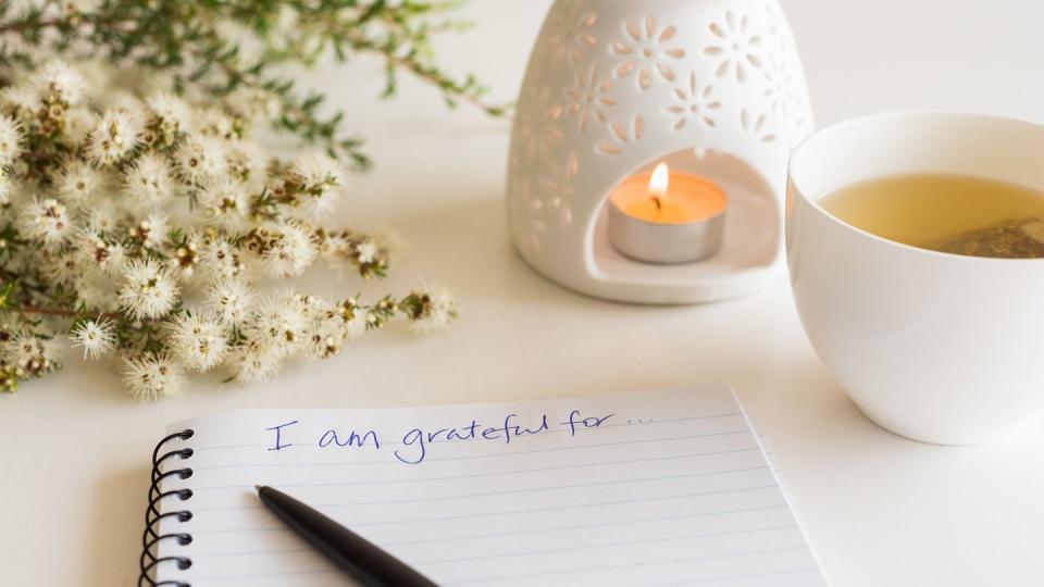 close up of handwritten text i am grateful for in foreground with notebook, pen, cup of tea, flowers and oil burner in soft focus