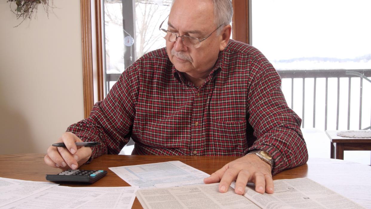 60s, Adult, Color Image, Document, Frowning, Gray Hair, Holding, Home Interior, Horizontal, Indoors, Looking Down, Male, Mature Adult, Men, Mustache, Objects, One Person, Only Man, Portrait, Preparation, Senior Adult, Senior Men, Serious, Table, Tax, calculator, casual, glasses, pen, sitting, tax form, window, working