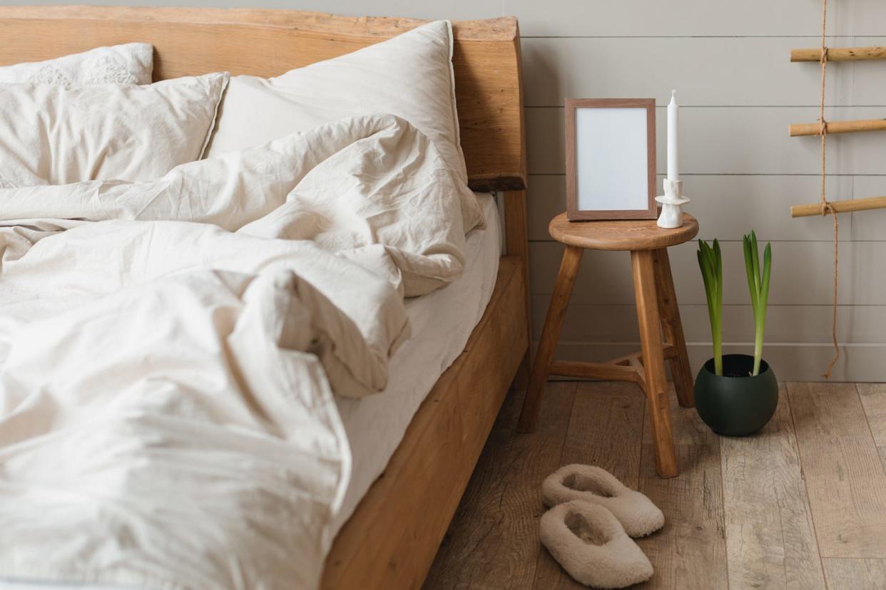 bedside table with a candle and blank picture frame, narcissus plant and pair of slippers next to an unmade bed in a bedroom